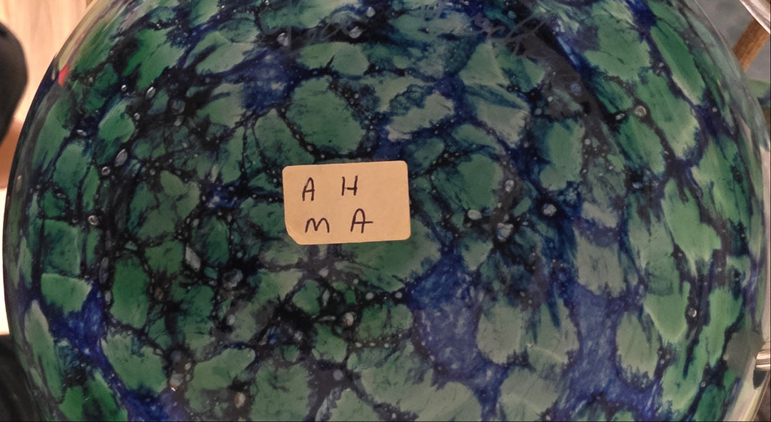A close-up of a round ceramic plate or bowl with a marbled green and blue glaze pattern, featuring a small handwritten paper label that reads &