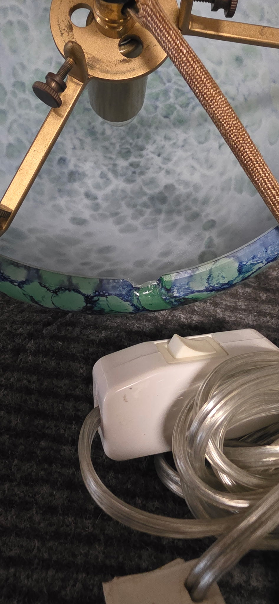 A close-up of a vintage-style table lamp base with a marbled blue and green glass pattern, a brass finial, and a white corded switch on a dark textured surface.