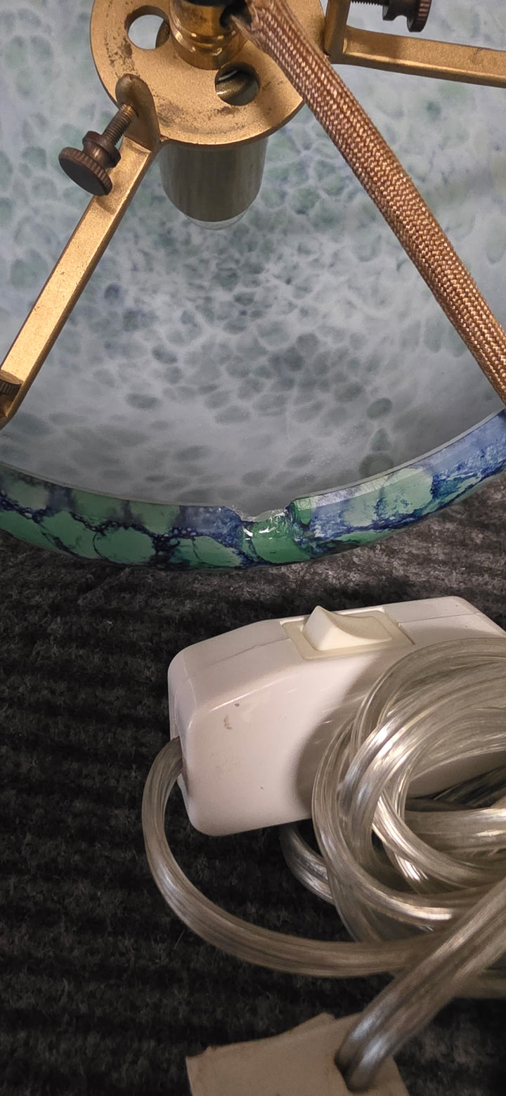 A close-up of a vintage-style table lamp base with a marbled blue and green glass pattern, a brass finial, and a white corded switch on a dark textured surface.