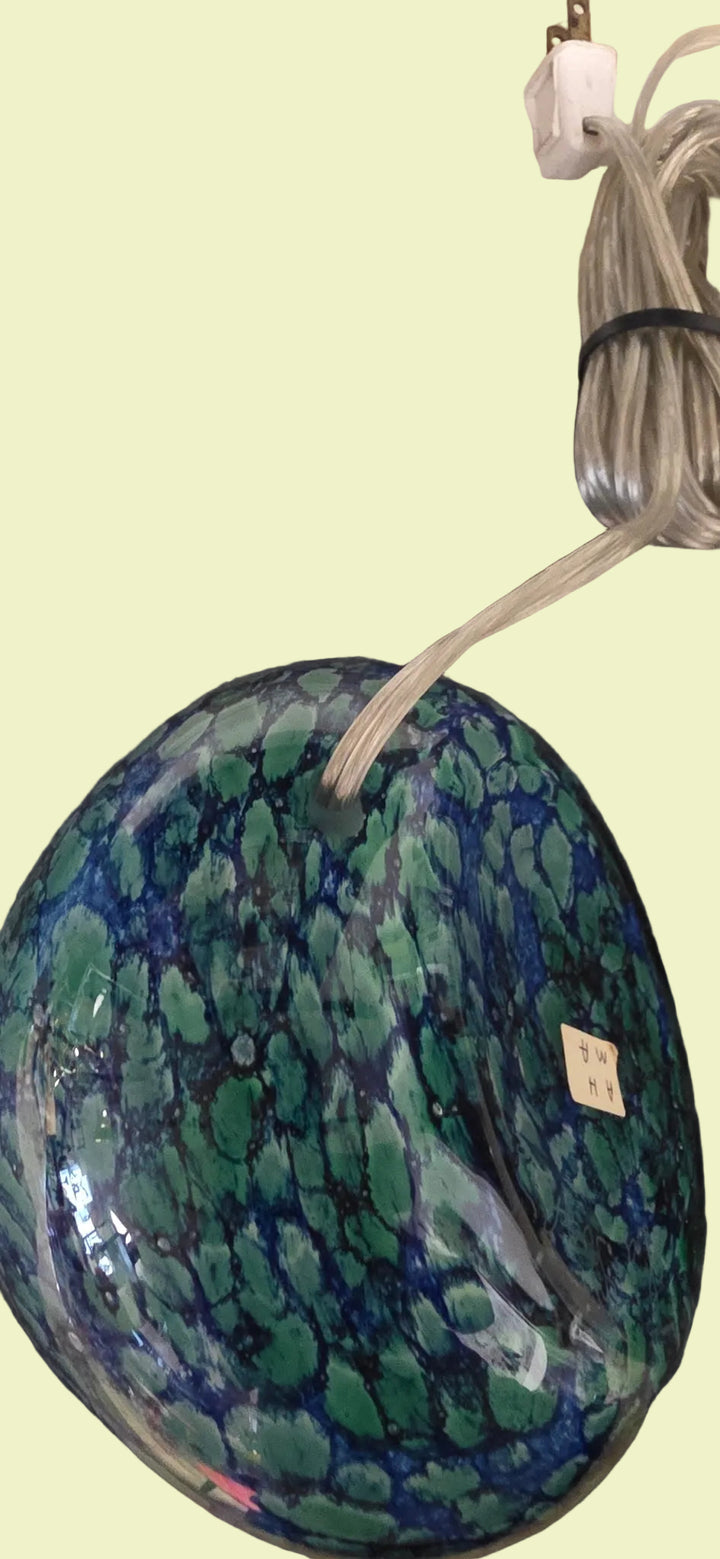 A large, round glass lamp base with a marbled pattern of green, blue, and black, resting on a dark gray textured rug with a coiled power cord visible.