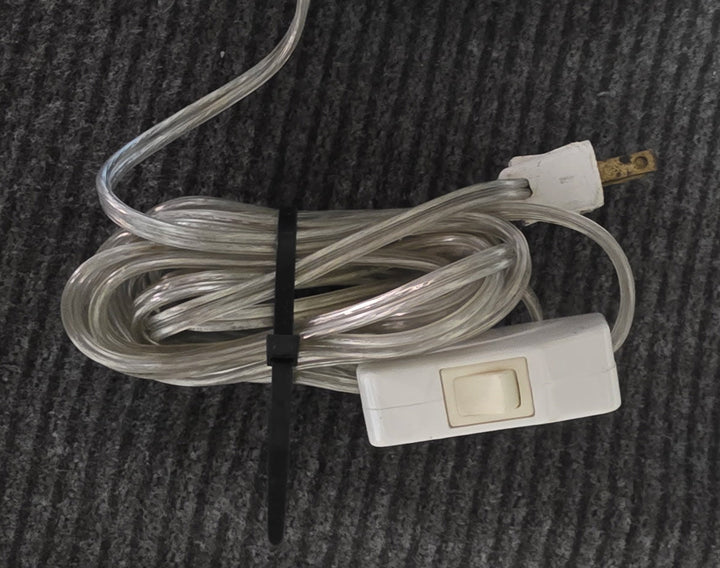 A coiled clear plastic cable with a white toggle switch connector and a black zip tie, placed on a dark gray ribbed fabric surface.