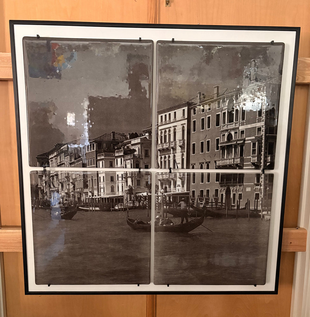 A large framed black and white vintage-style print depicting a canal scene with buildings and gondolas, displayed against a wooden door above a glass table.