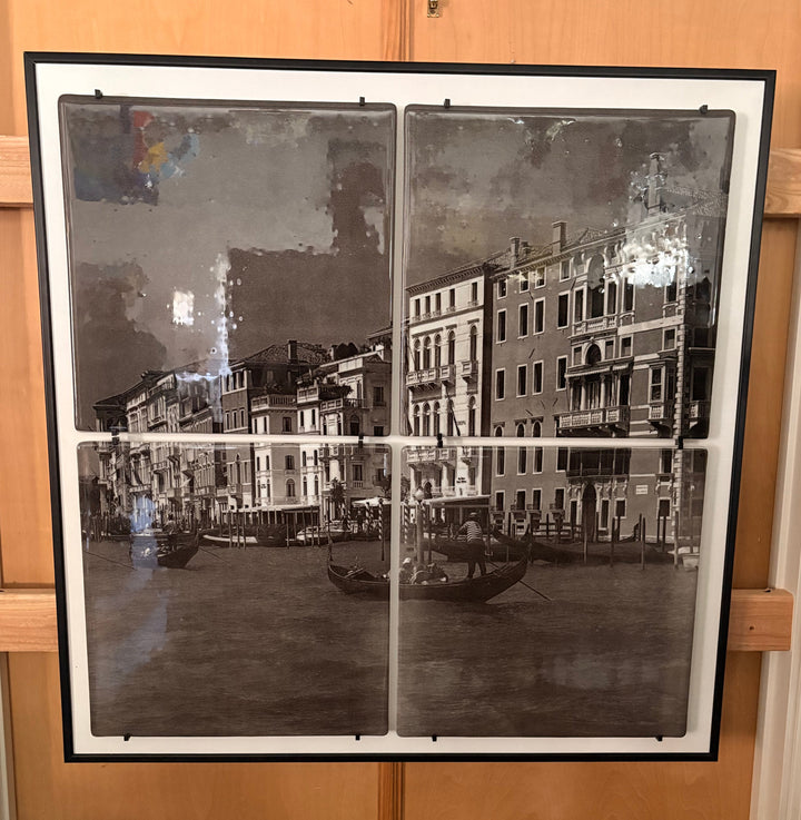 A large framed black and white vintage-style print depicting a canal scene with buildings and gondolas, displayed against a wooden door above a glass table.