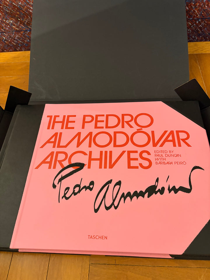 The Pedro Almodóvar Archives book by Taschen, featuring a bright pink cover with orange title text and a black autograph, displayed inside its black presentation box on a wooden floor.