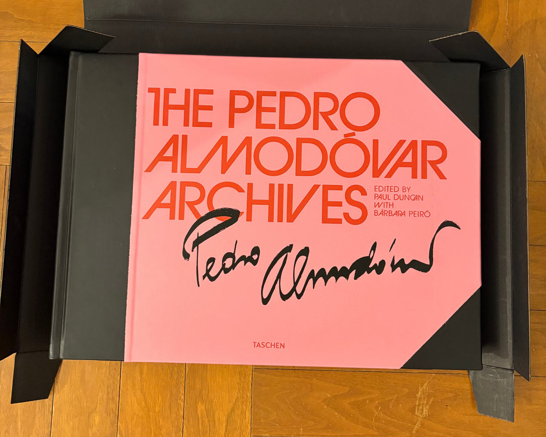 The Pedro Almodóvar Archives book by Taschen, featuring a pink cover with orange and black text, displayed inside an open black box on a wooden floor.
