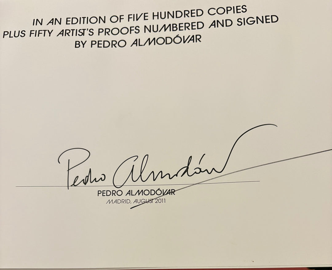 A close-up of a printed cover or page featuring black text about Pedro Almodóar signed copies, with a large signature and his name printed below, photographed over a wooden surface.