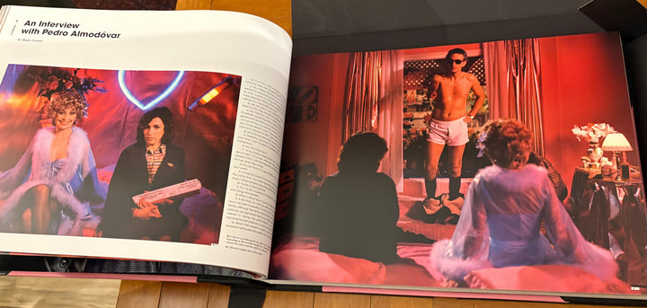 An open book or magazine spread photographed from above on a wooden floor. The left page shows a large red-toned photo with figures, and the right page shows printed text and a smaller matching photo.