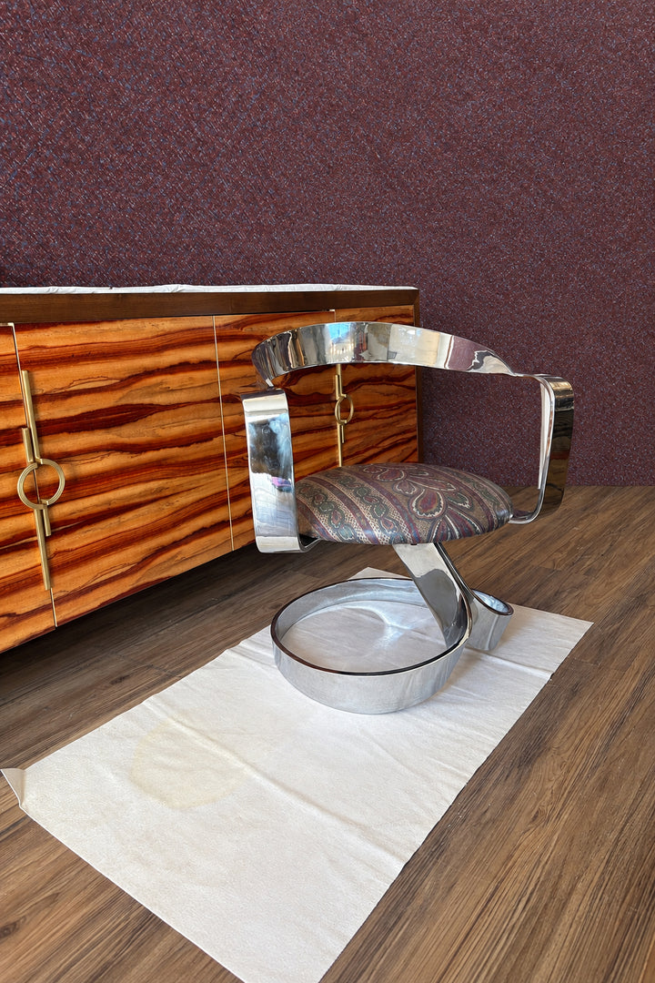 A mid-century modern chrome barber chair with a patterned upholstered seat, positioned on a white paper drop cloth on a wood floor in a showroom.