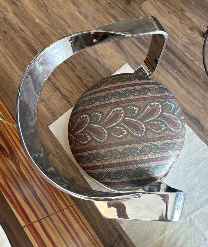 A modern curved chrome metal chair with a round upholstered seat cushion featuring a brown, green, and beige paisley pattern, shown on a wooden floor.