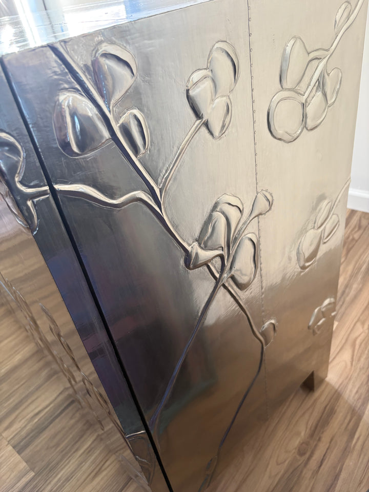 A close-up of a metallic silver cabinet or decorative panel with raised floral and leaf motifs, standing on a wood floor in an indoor room.