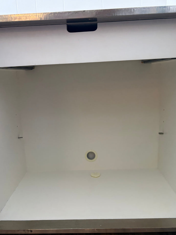 An empty white under-counter cabinet or storage compartment with a stainless steel countertop edge, interior walls, floor, and a small round drain opening on the back wall.