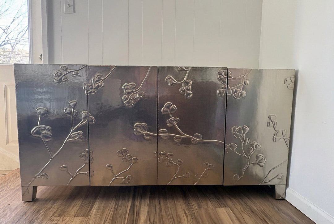 A modern five-door sideboard cabinet with a brushed silver metallic finish and raised floral branch patterns on the doors, shown in a room with wood floors and white paneled walls.