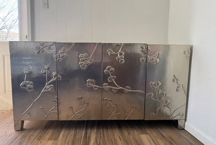 A modern five-door sideboard cabinet with a brushed silver metallic finish and raised floral branch patterns on the doors, shown in a room with wood floors and white paneled walls.