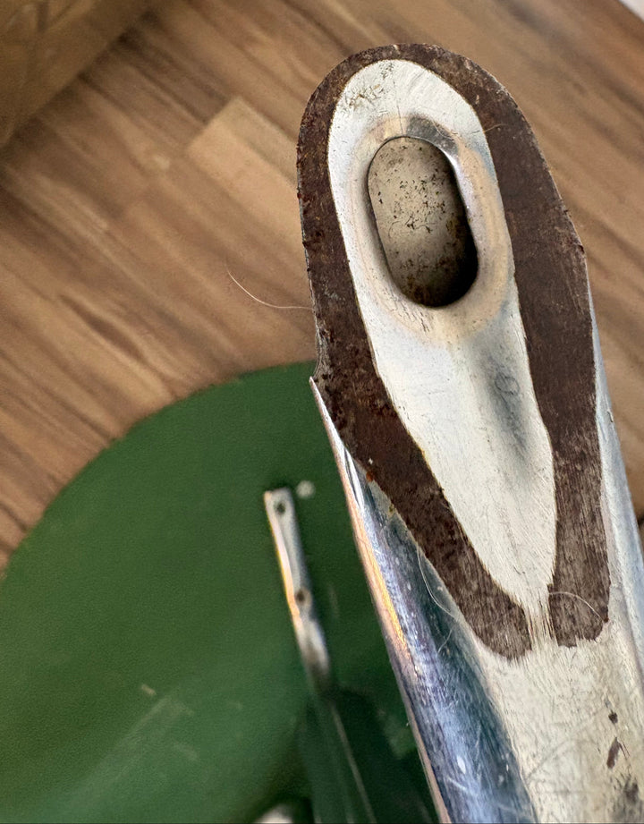 A close-up of a worn metal shaft or rod end with a dark brown rusted or oxidized layer around a light gray insert, photographed indoors on a wooden floor with a green circular object partially visible below.
