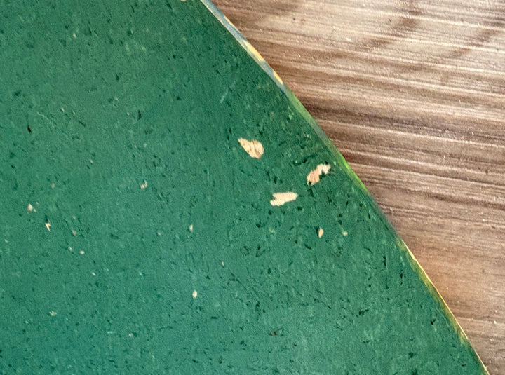 A close-up of a green textured material sample, likely a carpet or flooring sample, with small embedded flecks and a visible edge showing a tan backing, placed over a wood-grain surface.