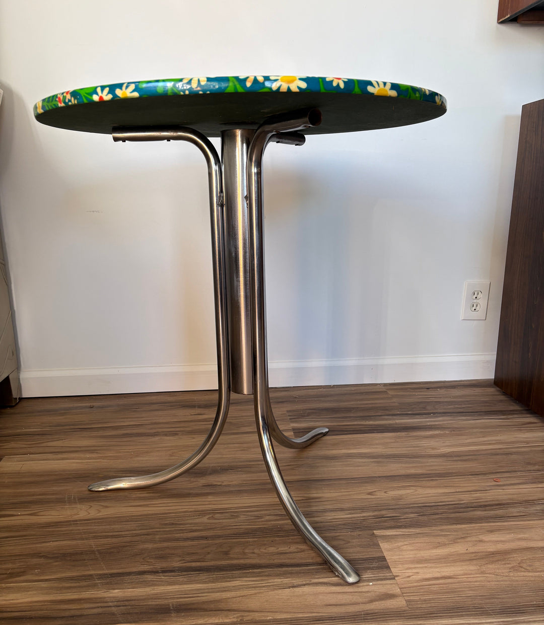 A mid-century modern tubular steel table with a green oval top featuring a yellow and white daisy floral pattern, photographed in a room with wood floors and a white wall.