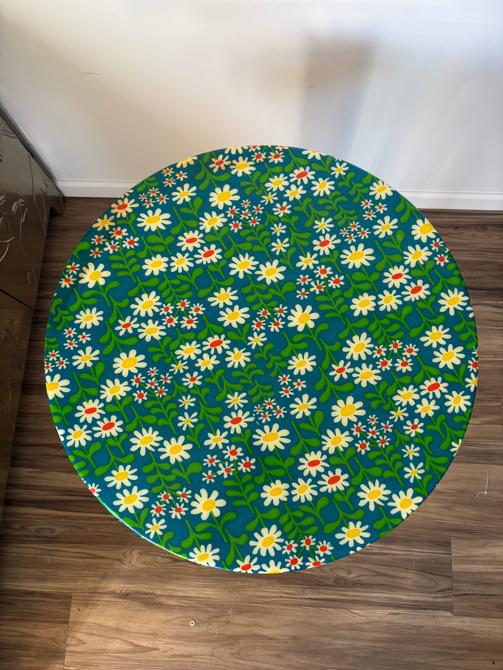 A round table covered in a vibrant floral print featuring white daisies with yellow centers, green leaves, and small red flowers on a dark teal background, placed on a brown wooden floor against a white wall.