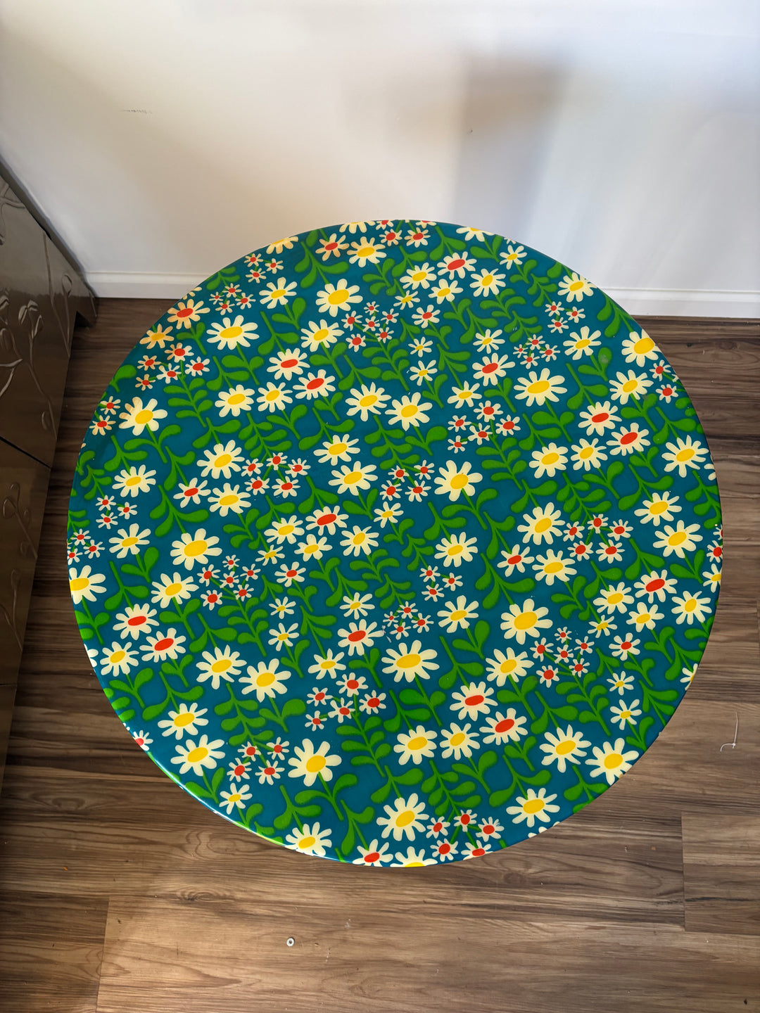 A round table covered in a vibrant floral print featuring white daisies with yellow centers, green leaves, and small red flowers on a dark teal background, placed on a brown wooden floor against a white wall.