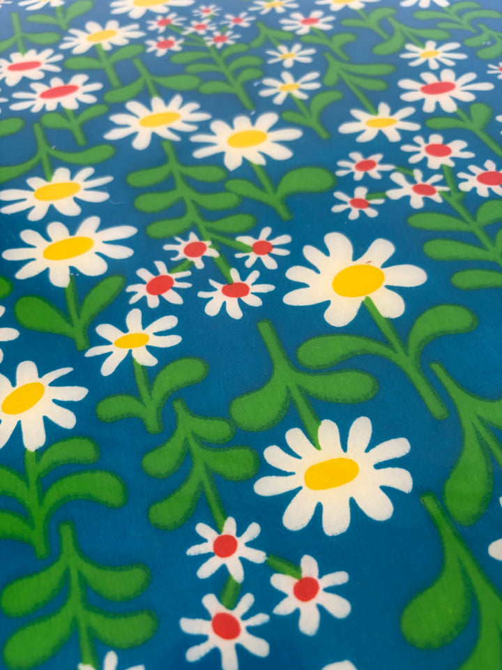 A close-up of a fabric or wallpaper pattern featuring white daisies with yellow centers and small red accents, set against a dark blue background with green leaves and stems.