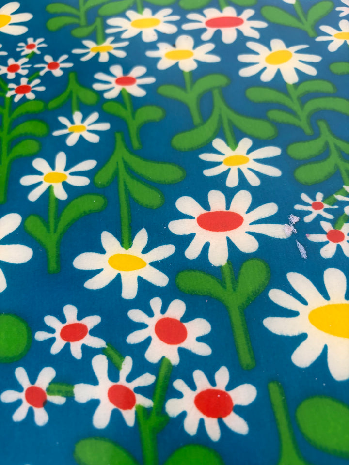 A close-up of a blue fabric or surface printed with a repeating pattern of white daisy flowers with yellow and red centers and green stems and leaves.
