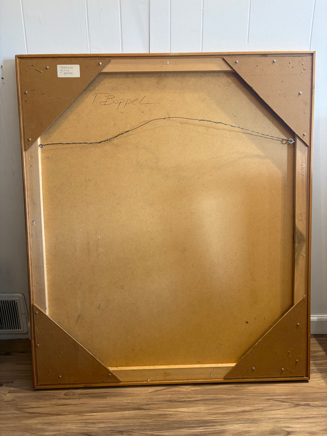 The back side of a large wooden picture frame leaning against a white paneled wall on a wood floor. The frame has a light brown wood finish with a tan cardboard backing and a visible hanging wire.