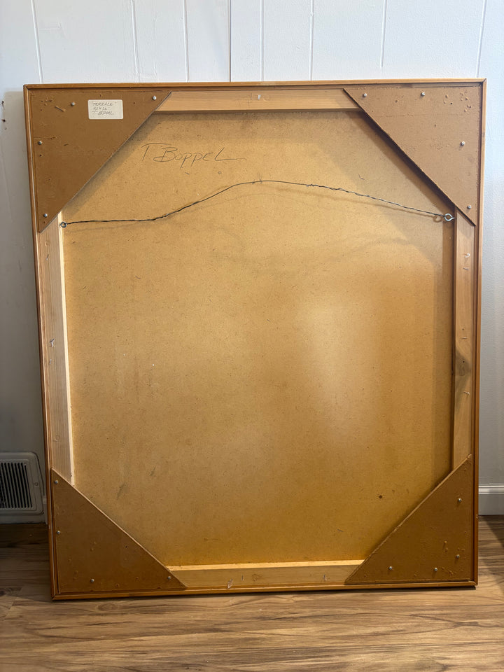 The back side of a large wooden picture frame leaning against a white paneled wall on a wood floor. The frame has a light brown wood finish with a tan cardboard backing and a visible hanging wire.
