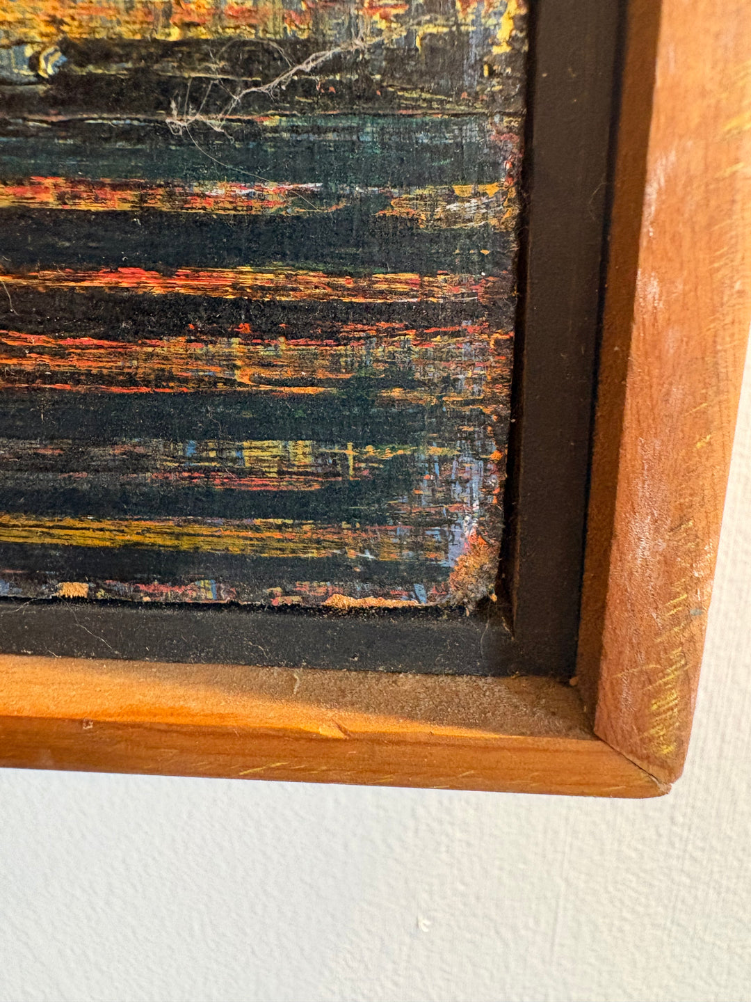 Close-up of a framed abstract painting showing textured horizontal brushstrokes in black, orange, yellow, and blue tones, with a warm brown wooden frame against a white wall.