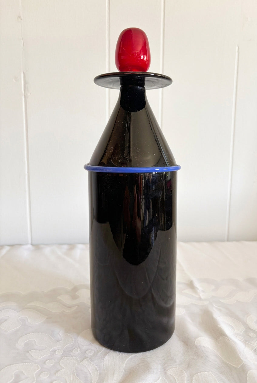 A glossy black decanter with a red cap and a blue band, on a white patterned fabric surface against a white paneled wall.