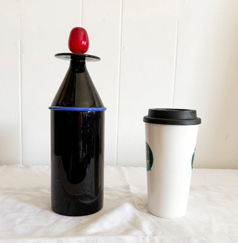 A black Murano glass decanter with a red stopper and blue band, shown alongside a white paper coffee cup on a white patterned fabric surface.