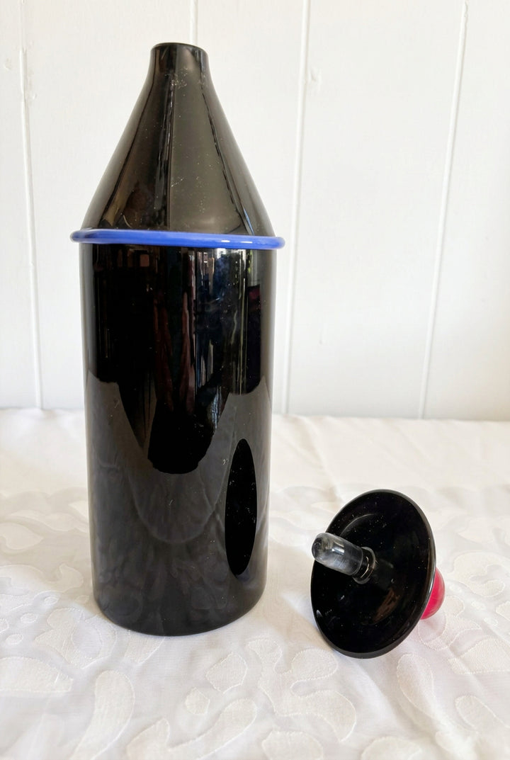 A glossy black Murano glass decanter or container with a pointed top section, shown alongside a detached black circular cap or lid with a small red top, placed on a white textured fabric surface against a white paneled wall.