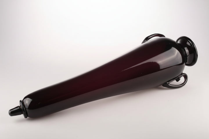 A dark, glossy glass bottle with a tapered body, a stopper on one end, and loop handles on the other, photographed on a neutral gray background.