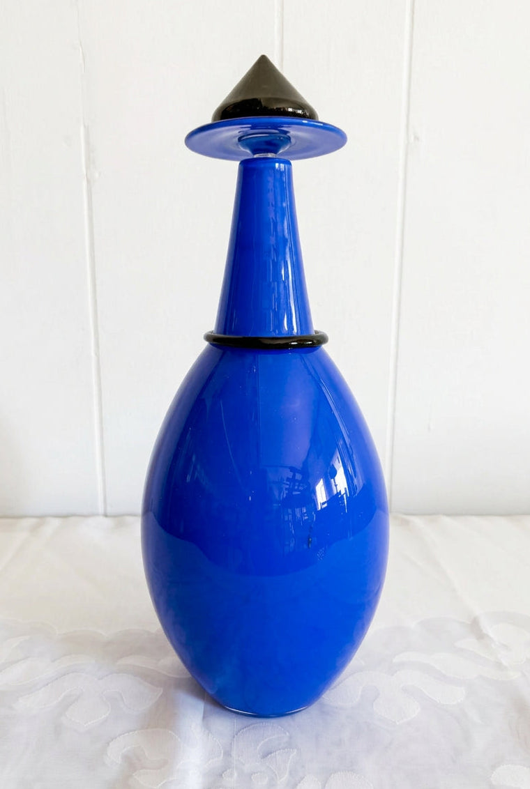 A cobalt blue glass bottle with a matching blue and black conical stopper, set on a white patterned tablecloth against a white paneled wall.