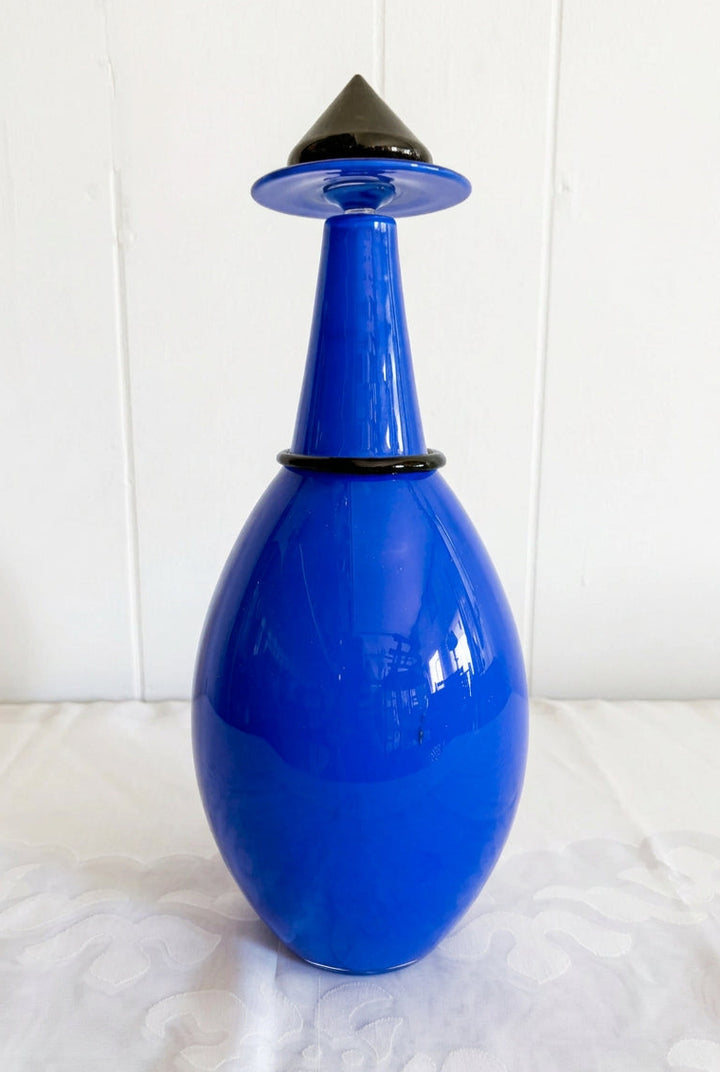 A cobalt blue glass bottle with a matching blue and black conical stopper, set on a white patterned tablecloth against a white paneled wall.