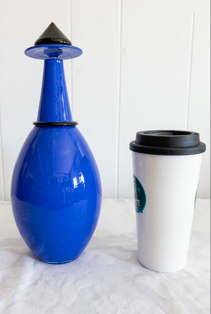 A blue glass decanter with a black conical cap and a white travel tumbler with a black lid, both resting against a white paneled wall and textured white fabric.
